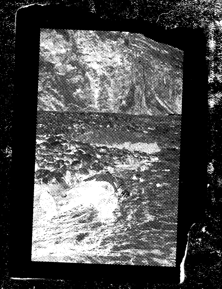 Alt text: Inverted picture of a torn piece of paper with a halftone image on it lying on a stone like background. Halftone image shows a Tyler lying in a shallow stream next to rocks. The way he is lying makes him appear to be almost dead. Because of the angle, only the lower half of his body is visible and not his face. A cliff can be seen in the background. Tyler is wearing black pants and a lighter colored shirt with black boots. This is the same setting and outfit as the Jumpsuit music video.