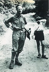 Alt text: Uncropped black and white image of child not edited in halftone. Child shown standing next to adult man with short dark hear wearing dark long sleeved shirt, dark pants, black boots, and glasses. Man has jacket hanging from one arm and both hands on hips. Both are standing on a dirt road and trees can be seen in background.